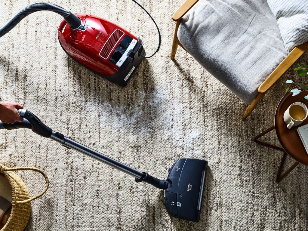 A person is vacuuming a textured carpet in a living room with a red vacuum cleaner, near a wooden chair and small table.